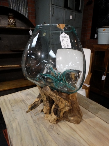 Clear, rounded glass vase perched on a twisted wooden stand, placed on a wooden table in a store. A tag hangs from the rim.