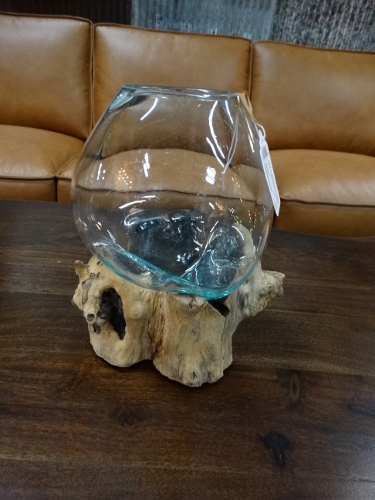 Clear glass fishbowl resting on a driftwood stand atop a dark wooden coffee table, with a tan leather couch in the background.