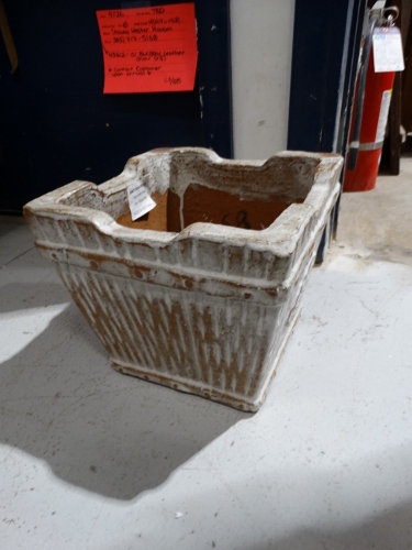 Rectangular, weathered concrete planter with soil inside sits on a concrete workshop floor. A red note is posted on the wall behind it.